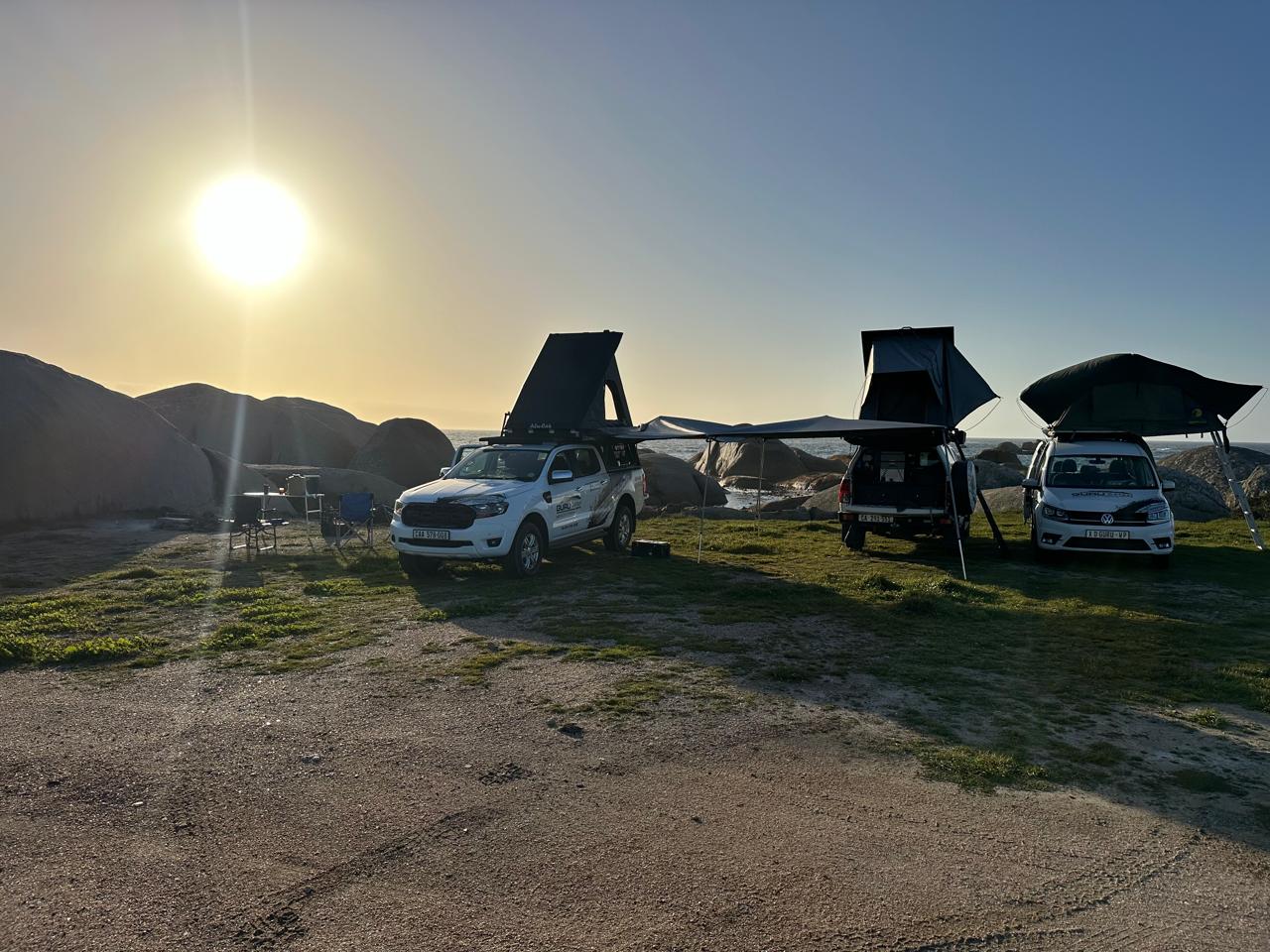 Two white Guru Safari rental vehicles, a Ford Ranger 4x4 and a VW Caddy camper, are parked side-by-side with their rooftop tents and awnings deployed, overlooking the ocean at sunset with large coastal boulders in the background. Camping chairs are set up nearby.