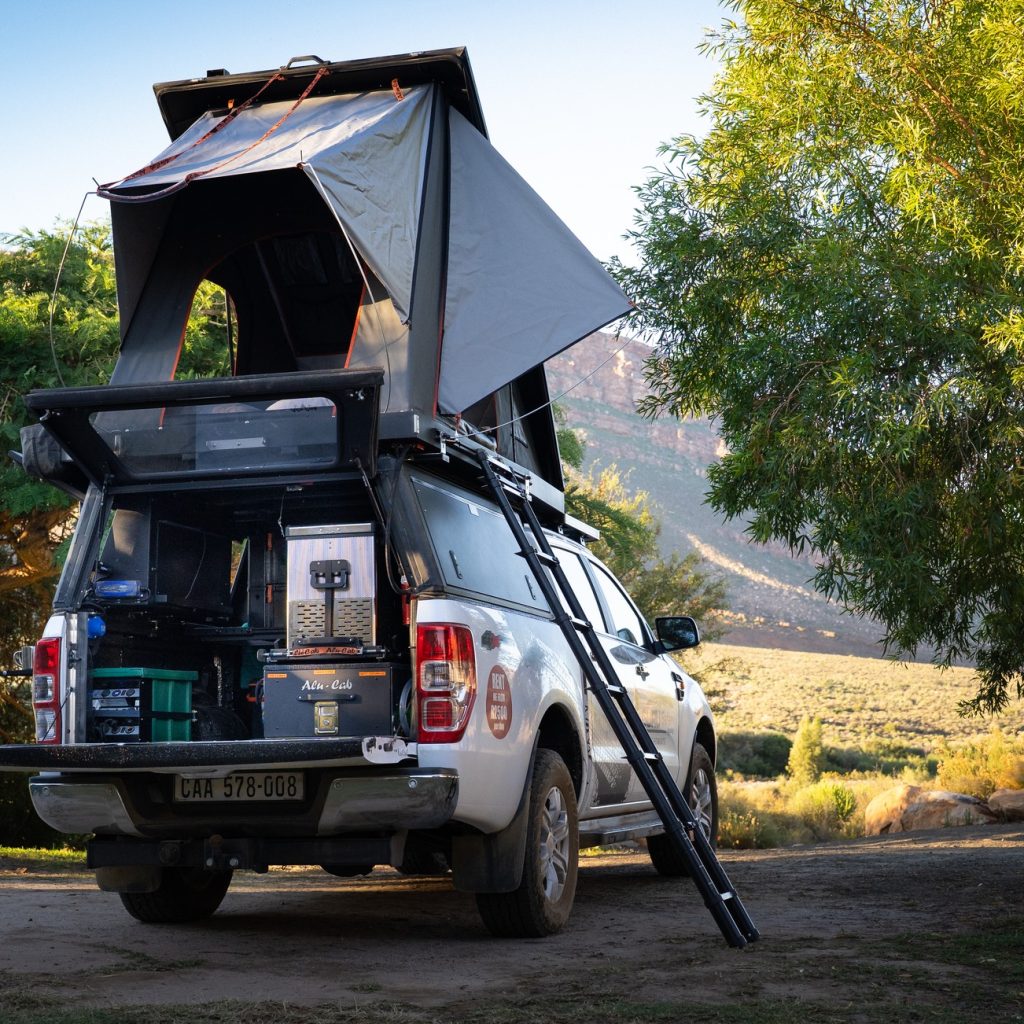 Rear view of a white Ford Ranger 4x4 rental truck (bakkie) parked at a campsite, featuring an open hard-shell rooftop tent, a deployable ladder, and the tailgate open to show a fitted camping kitchen, fridge, and storage system.